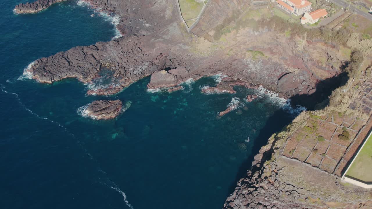 Stunning aerial footage of São Miguel Island's rugged volcanic coastline, featuring dramatic black rocks, deep blue waters, and vibrant orange rooftops under clear skies