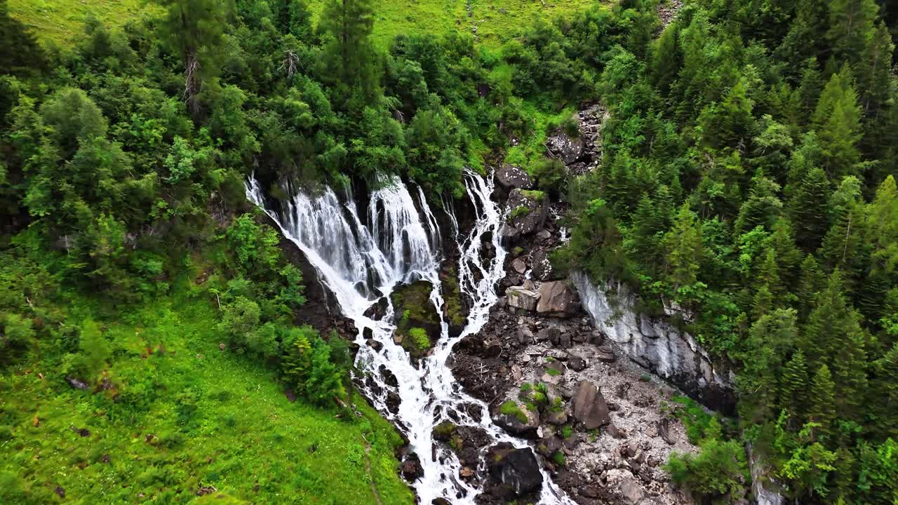 Cascata di Sibenfaelle che scende a cascata lungo un fianco montuoso scosceso, avvolta da fitte pinete e abbondante vegetazione in una giornata nuvolosa a Lenk, Svizzera, drone che si avvicina