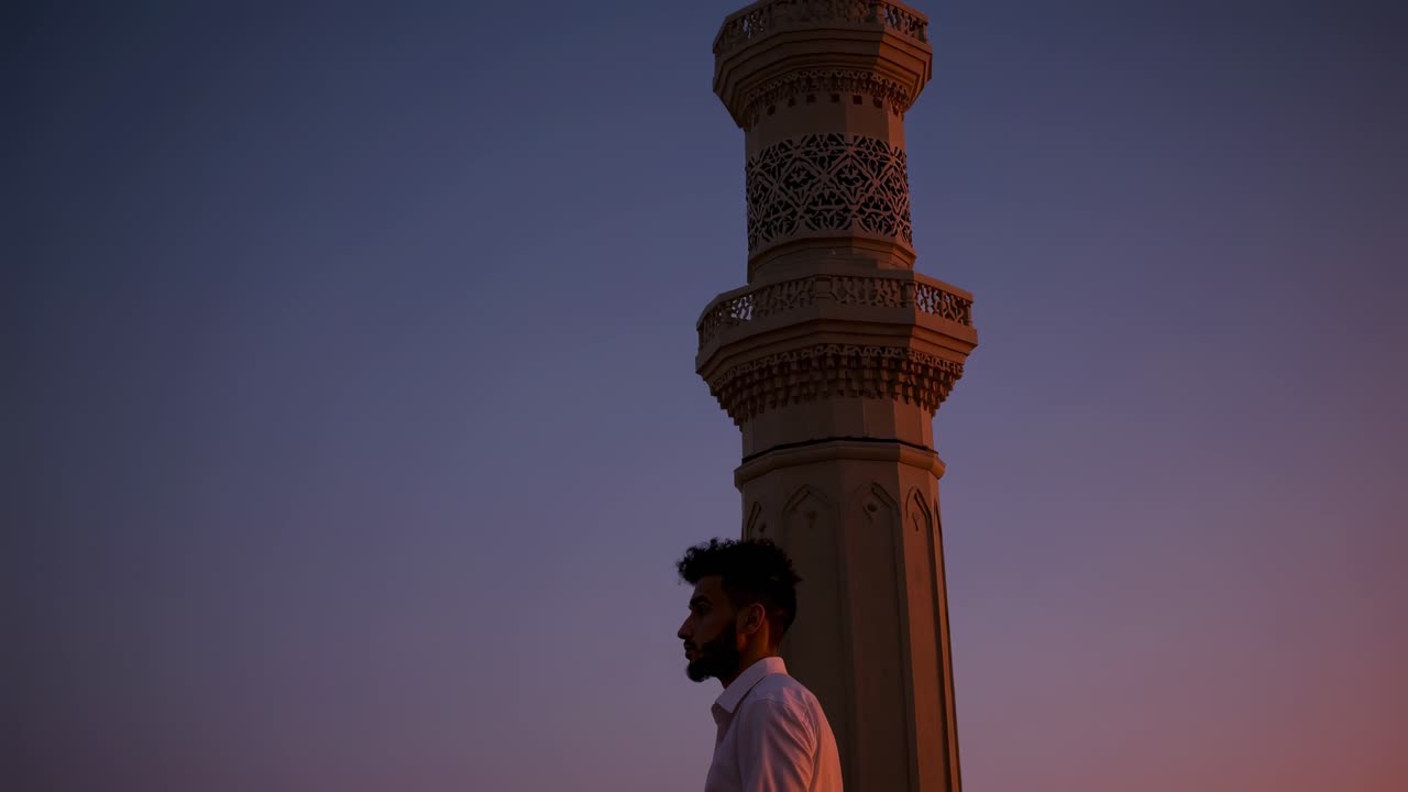 Young bearded Muslim man in a white shirt praying near a minaret during a vibrant sunset, enveloped in a serene and spiritual atmosphere that evokes deep contemplation and devotion