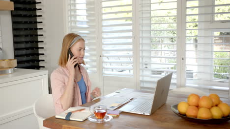 working from home, woman using laptop and talking on smartphone in kitchen