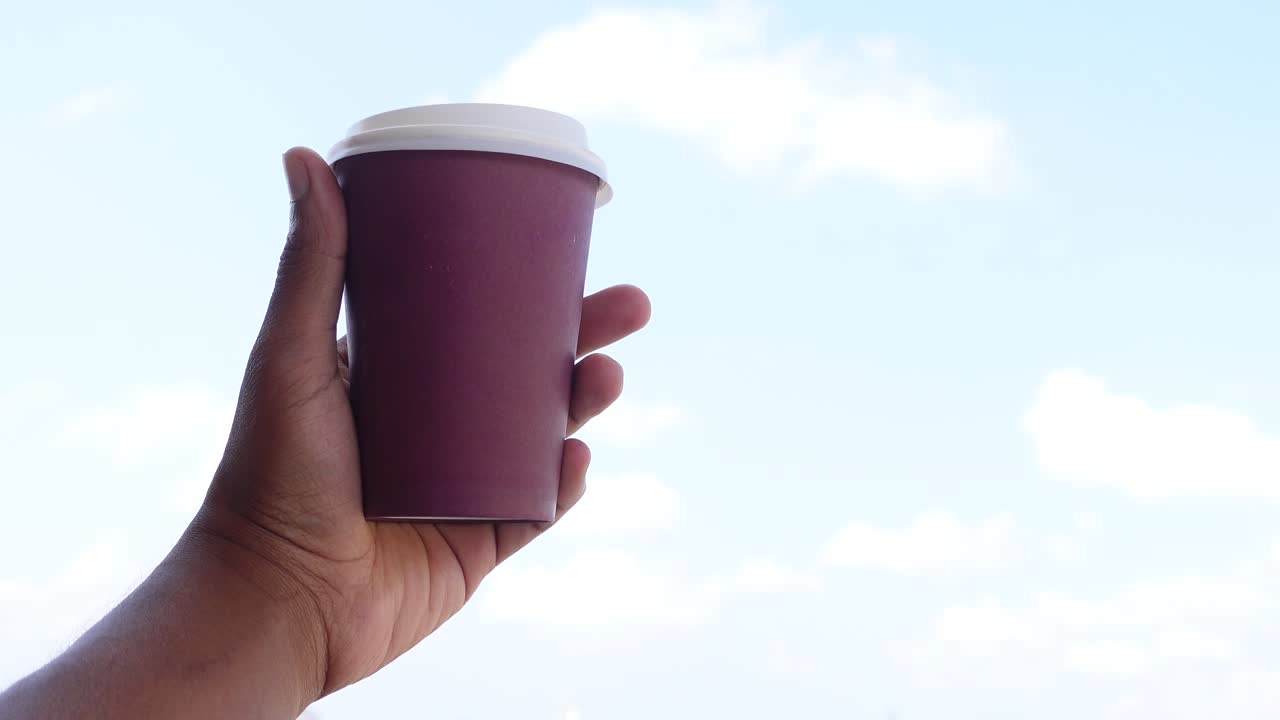 mano sosteniendo una taza de café de papel púrpura contra un cielo nublado