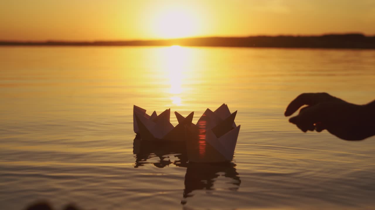 Female's hand putting one paper boat on the water and it joins to other ones at sunset. New origami ship adding to the rest during beautiful sunset with the sun reflection in the lake