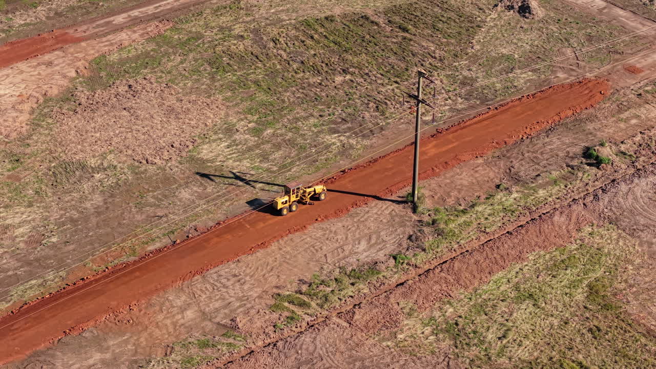 Aerial view follows lone bulldozer moving beside power grid tower, with dust swirling in afternoon sun. Ideal for infrastructure or heavy machinery content.