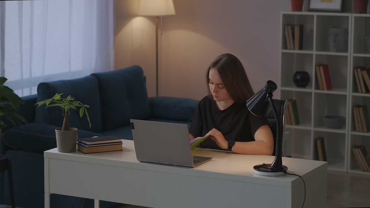 저녁에 집에서 멀리서 일하는 젊은 여성은 노트북을 가지고 일하고 현대 아파트의 거실에서 테이블에 앉아 노트북에 메모를 하고 있습니다.