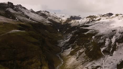 Aerial-view-of-a-snow-capped-mountain-gorge-at-Sólheimajökull,-showcasing-rugged-terrain-and-a-winding-glacier-river