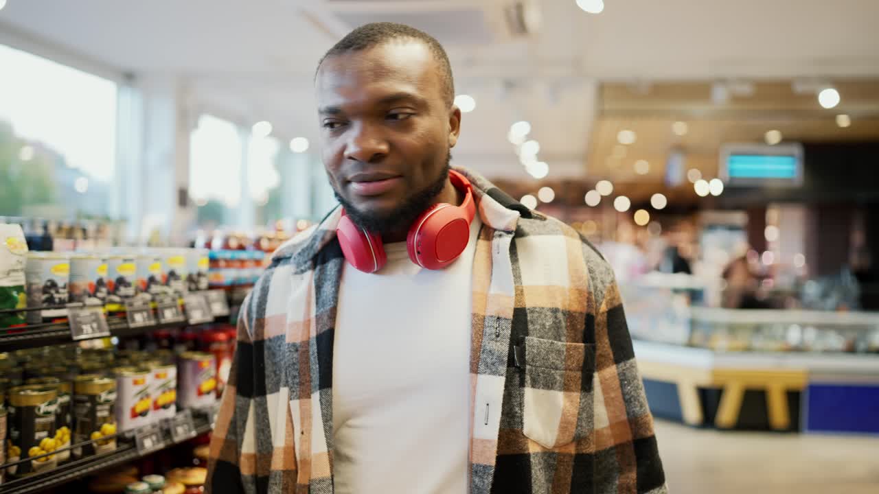 A confident and happy man with Black skin in a checkered shirt and red wireless headphones looks at the counters and looks at the necessary goods on modern shelves in a large grocery supermarket
