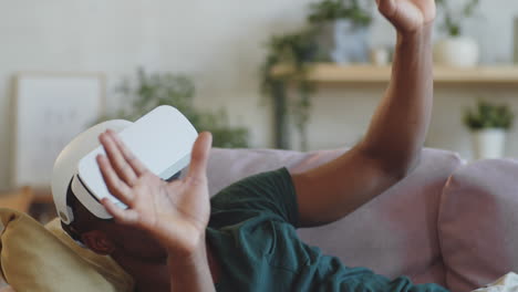 Delightful-Black-Man-Using-VR-Headset-while-Lying-on-Sofa