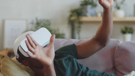 hombre negro encantador usando auriculares vr mientras está acostado en el sofá