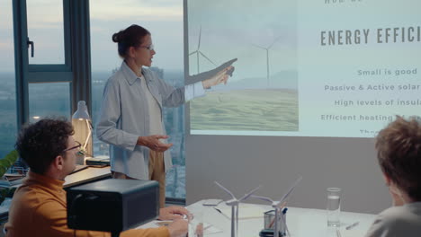 mujer de negocios de pie junto a la pantalla de proyección y dando una presentación a sus colegas sobre la eficiencia energética durante una reunión de oficina en una compañía de energía verde