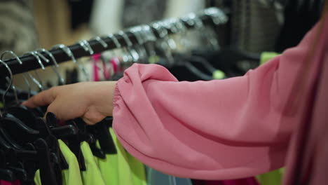 hand in pink shirt reaching for a lemon green tight from a clothes rack in a store, other clothing items are visible in the background as she observes the garment closely, with a blur background