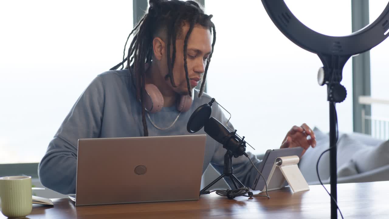 Mid adult African American man working laptop at home desk adjusting tablet and tapping for podcast