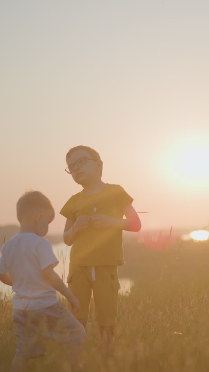 Woman in a blue gown, using a headset and smartphone, stands with two boys in a sunlit field at sunset, as one boy in white approaches
