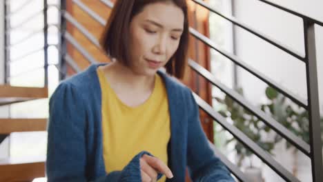 Video-of-happy-asian-woman-smiling,-sitting-on-stairs-using-tablet-and-looking-around-at-home