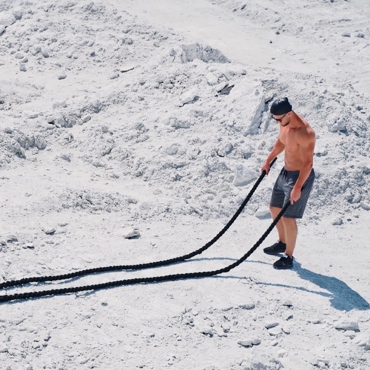 fitness athlete in a cap and sunglasses prepares for training with ropes outdoor. Heavy training in a quarry. Intense workout with ropes in white mountains. Strong attractive bodybuilder. Lifestyle. White landscape.
