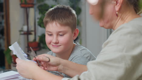 woman cutting paper into shapes with scissors while boy in grey top watches and smiles, crafting together at table surrounded by paper scraps and greenery in cozy bright indoor environment