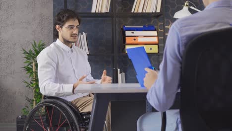 se sienta en el lugar de trabajo en una silla de ruedas, comparte ideas creativas y puntos de vista con el trabajador masculino.