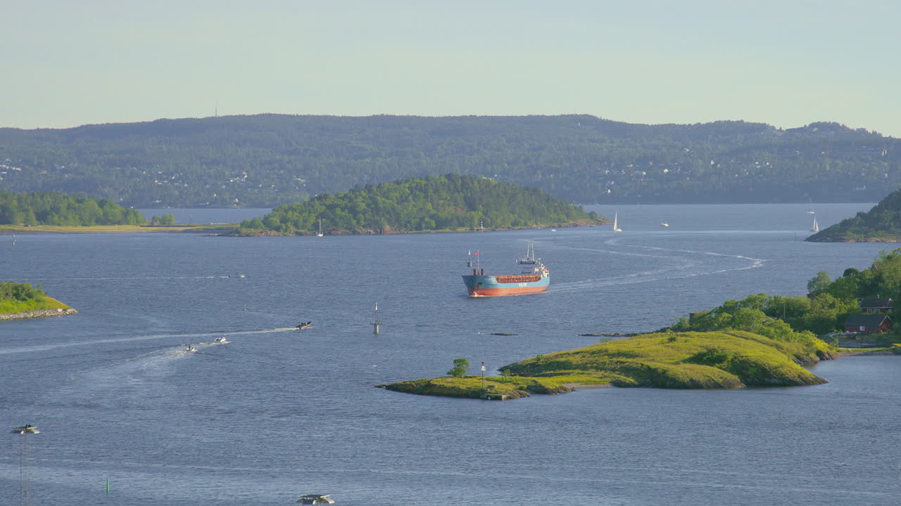 Cargo ship and sailboats on calm waters surrounded by lush green islands under a clear sky.
