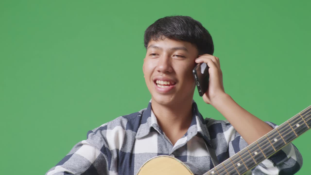 초록색 화면 배경 에 서 있는 동안 기타 를 들고 스마트폰 에 대화 하는 아시아 청소년 의 근접 사진