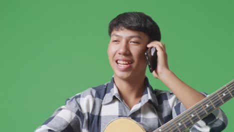 close up of asian teenager with guitar talking on smartphone while standing on green screen background