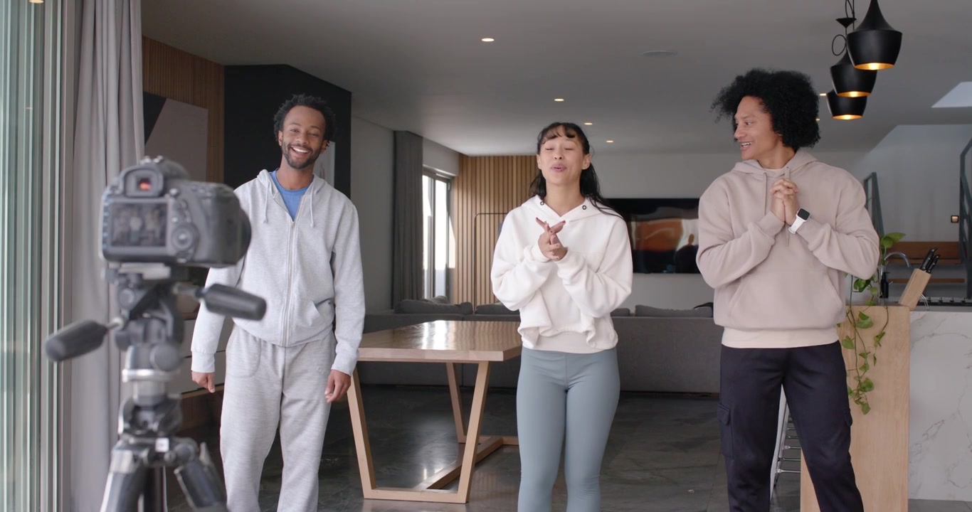 Diverse friends in hoodies waving clapping as camera on tripod with mic recording greeting at home
