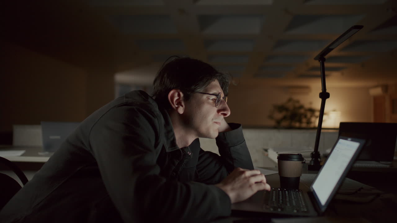 Young Man Reading Text on Laptop as Working Overtime in Office at Night
