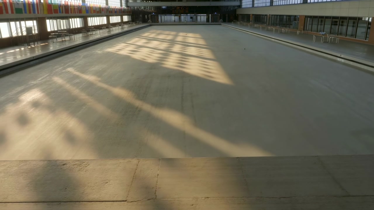 Wide interior view of a vacant swimming pool basin with smooth concrete surfaces, sharp diagonal light and shadow shapes across the floor, and modern walls