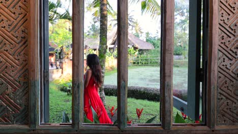 Mujer-Asiática-Vestida-De-Rojo-Caminando-Por-Una-Ventana-De-Madera-Tradicional-En-Un-Día-Soleado-En-Bali,-Indonesia