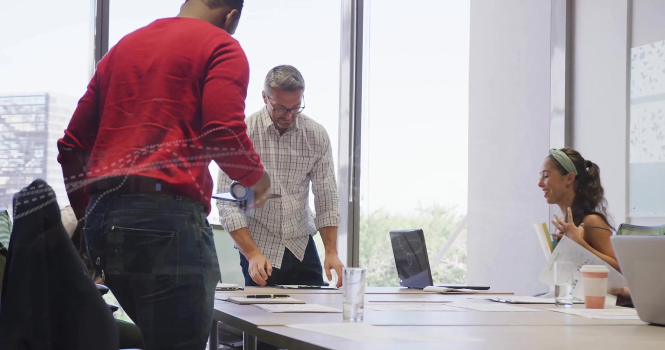 Equipo levantándose de las sillas y reuniéndose en la mesa mientras un presentador con suéter rojo revisa la tecnología de cascos de realidad virtual.