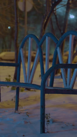 banco cubierto de nieve se encuentra solo en el anochecer de invierno, iluminado por la luz ambiental suave, los árboles circundantes y el fondo borroso de los elementos urbanos