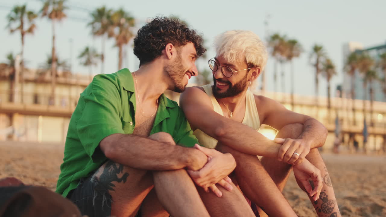 Pareja gay feliz sentada y hablando en una playa al atardecer