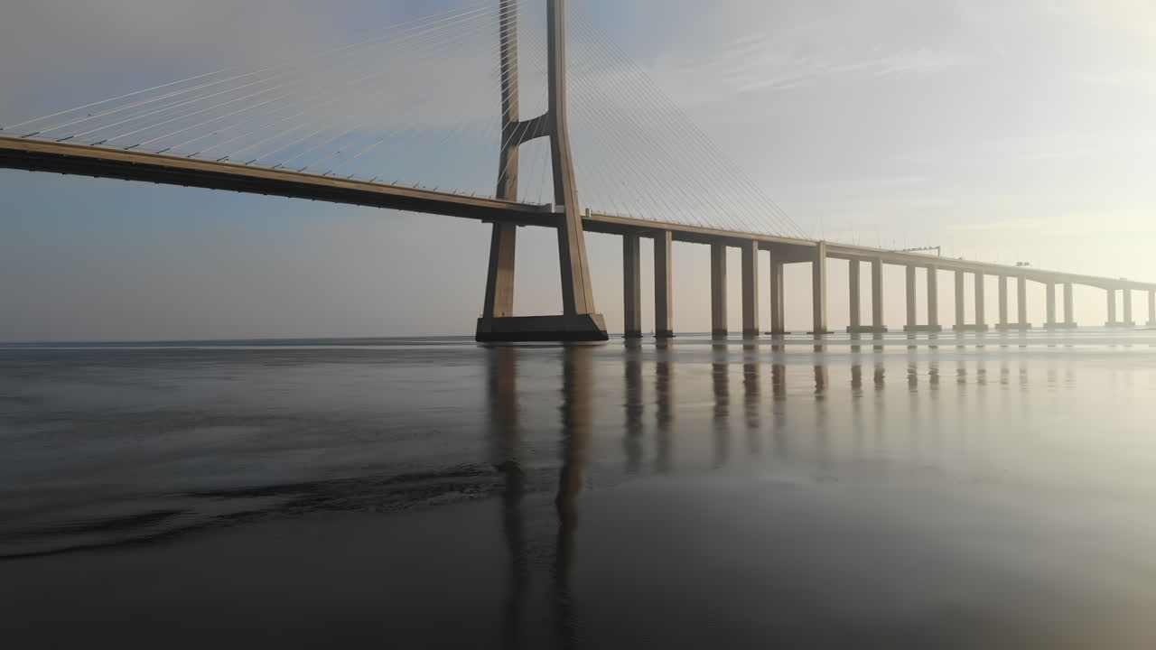 A breathtaking view of a massive bridge standing over serene water at dawn. The calm surface reflects the structure, showcasing its impressive design in soft morning light