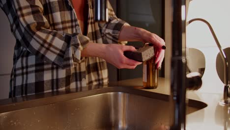 A-confident-brunette-girl-in-a-checkered-shirt-takes-out-a-gray-dishwashing-sponge-pours-a-special-detergent-on-it-and-washes-the-dishes-in-the-evening-during-her-routine-in-a-modern-apartment