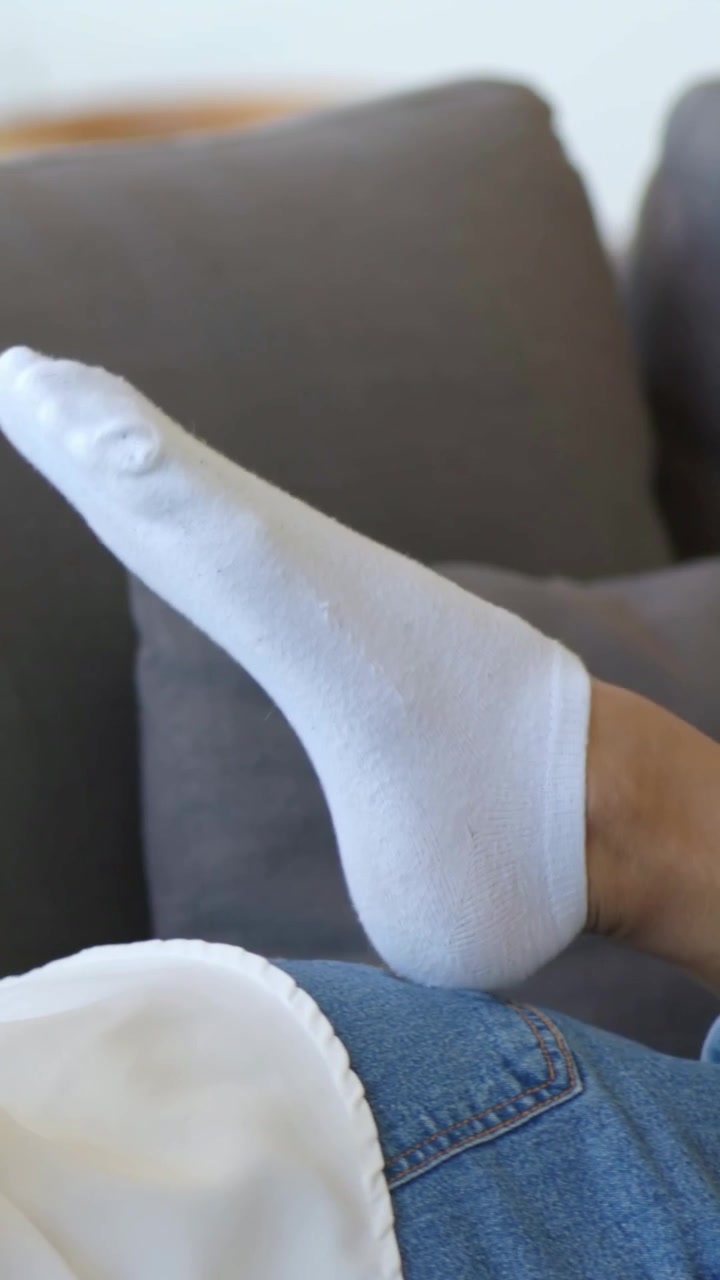 Video verticale: Donna sdraiata con jeans sfrangiati che muove i piedi con calzini bianchi su un divano grigio, sistemandosi.