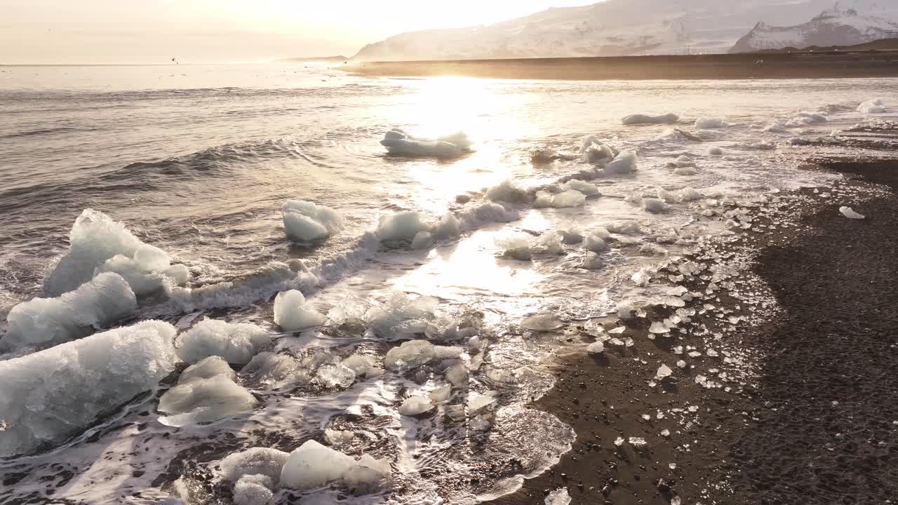 Drone flying backward revealing scattered ice chunks on the shore of Diamond Beach during a golden sunrise in Jökulsárlón, Iceland.