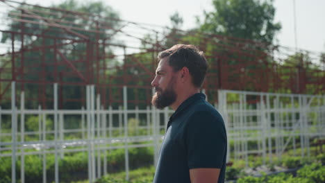 man turning head looking at camera while standing near metal frame and green crops on sunny farm day, side profile showing beard and blue shirt, background