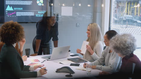 Vista-A-Través-Del-Cristal-De-Mujeres-Empresarias-Que-Colaboran-En-Una-Reunión-Creativa-Alrededor-De-Una-Mesa-En-Una-Oficina-Moderna