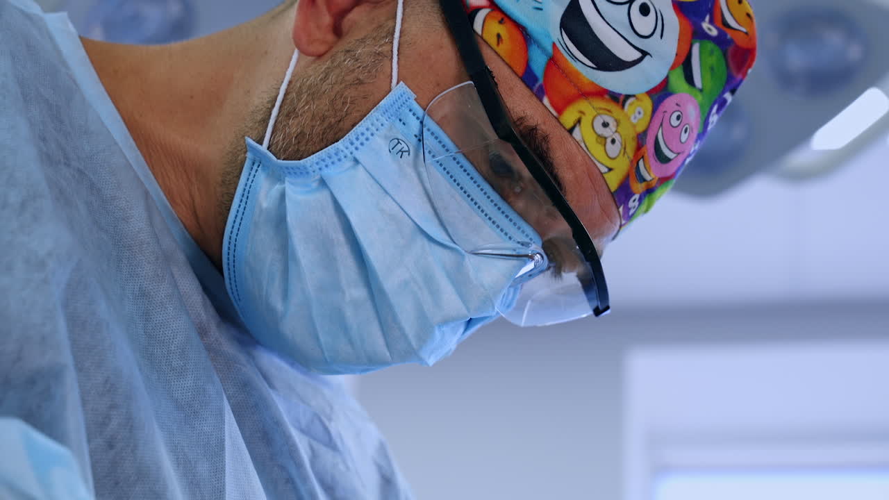 Male bearded surgeon wearing funny colorful cap, mask and glasses. Portrait of a professional focused on procedure. Close up.