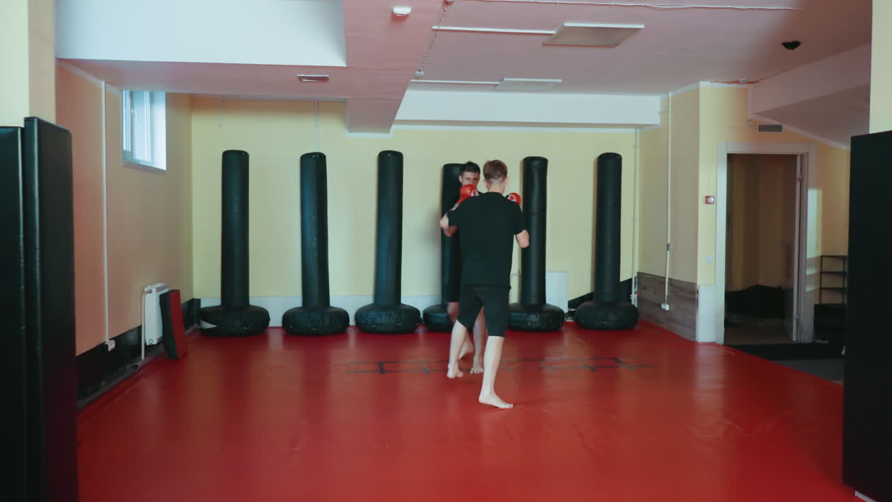 Atletas entrenando en un gimnasio de artes marciales sobre una colchoneta roja; un atleta con guantes rojos lanza un puñetazo extendido hacia un oponente con guantes negros, ambos descalzos demostrando concentración y fuerza en el entrenamiento de combate.