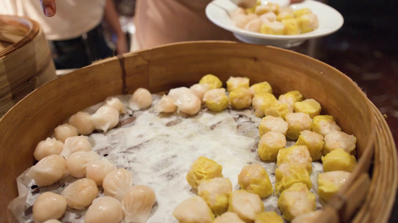 A person uses metal tongs to select dim sum from a large bamboo steamer basket