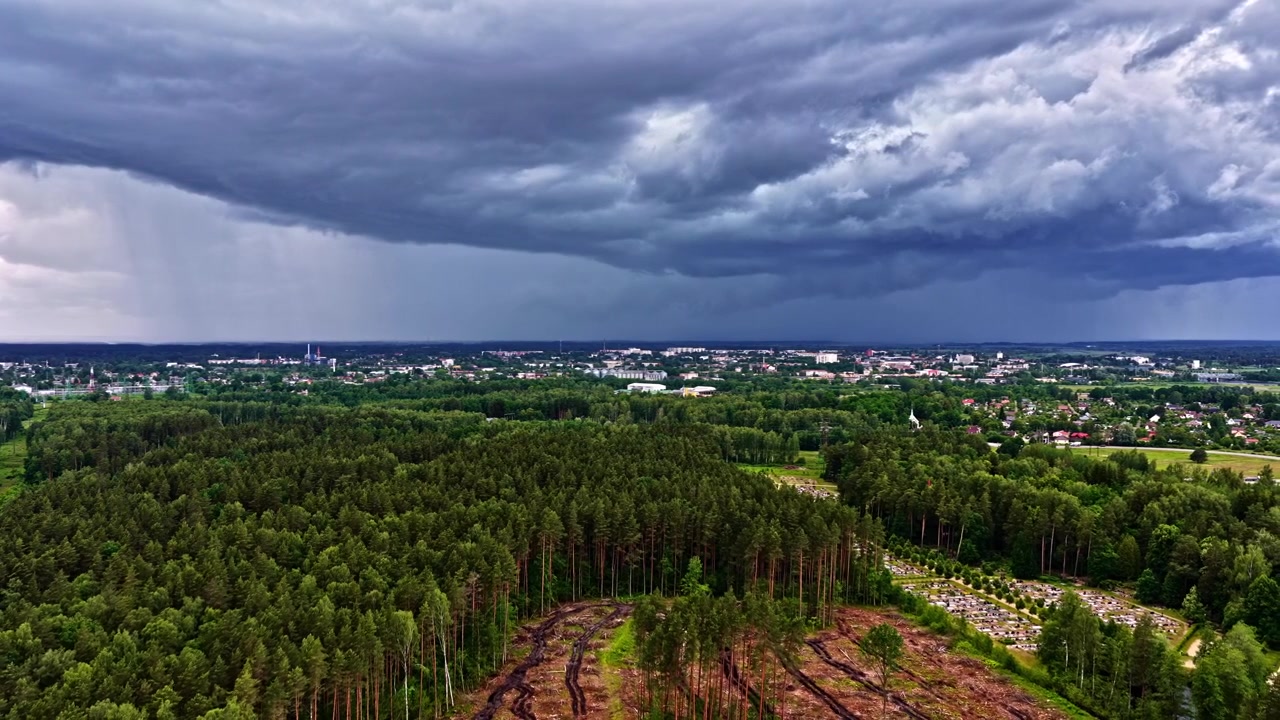 Eine weite Drohnenaufnahme, die dunkle, schwere Sturmwolken über einem üppig grünen Wald und einer Stadt einfängt