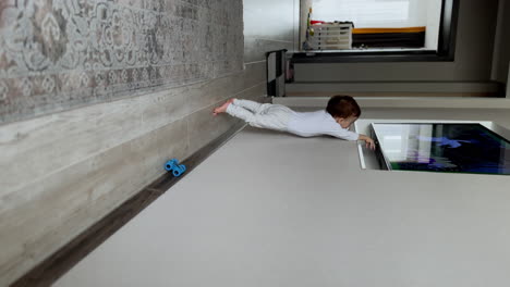 caucasian baby boy in white clothes walking by the room holding a candy. little child tries to reach a tv on the wall. vertical screen.
