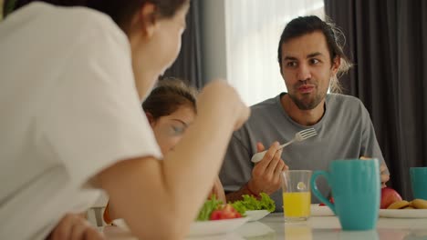 Close-up-shot:-happy-brunette-man-eats-salad-during-his-breakfast-together-with-his-little-daughter-in-a-yellow-dress-from-his-wife-in-a-white-T-shirt-at-a-white-table-in-a-modern-apartment-in-the-morning