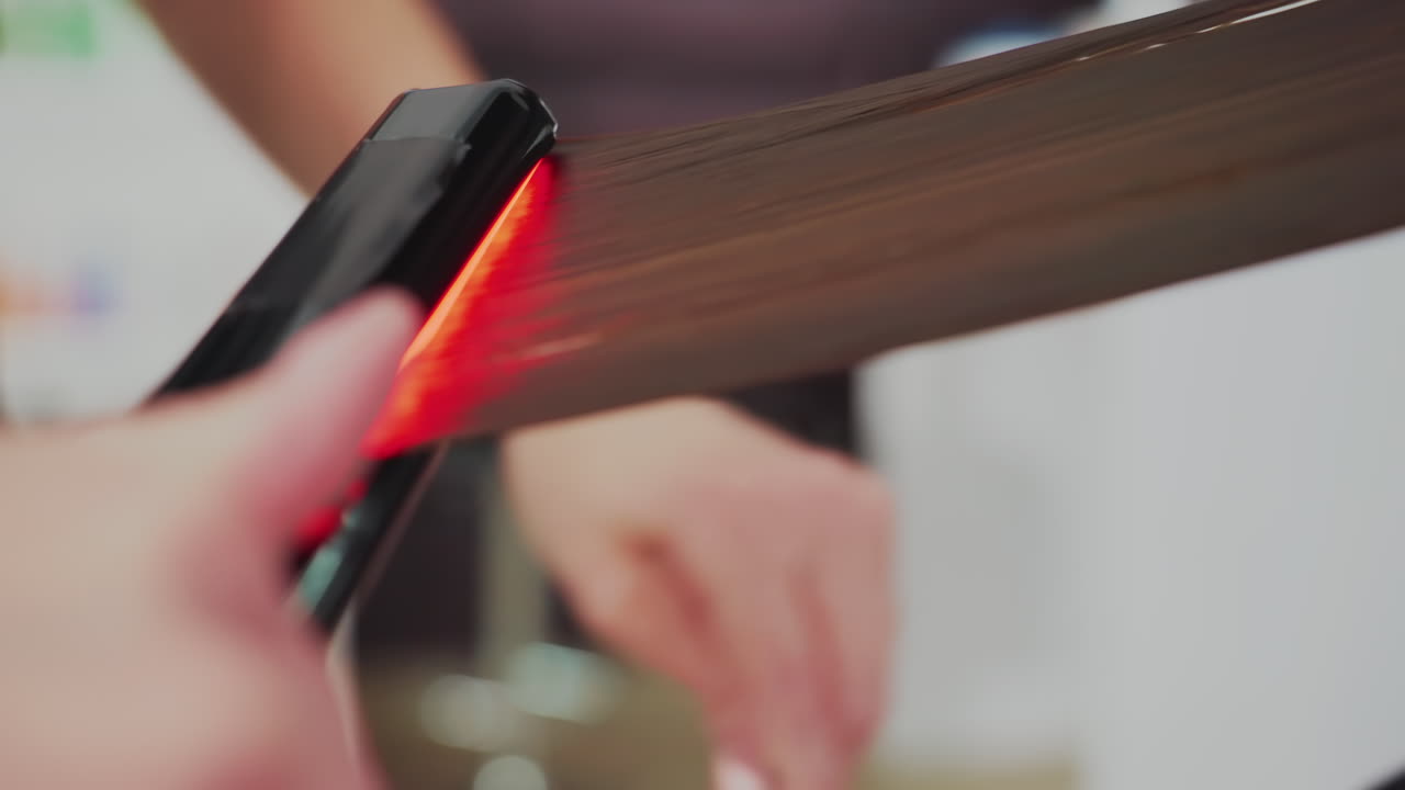 클로즈업 장면: 흰색 머리카락을 따라 미끄러지듯 움직이는 납작한 고데, 스타일리스트가 열판으로 머리 섹션을 고정하고 매끄럽게 다듬을 때 여성 고객이 열 교정 시술을 받고 있으며, 빨간색 열 표시등과 안정적인 손길은 정확성을 강조합니다.