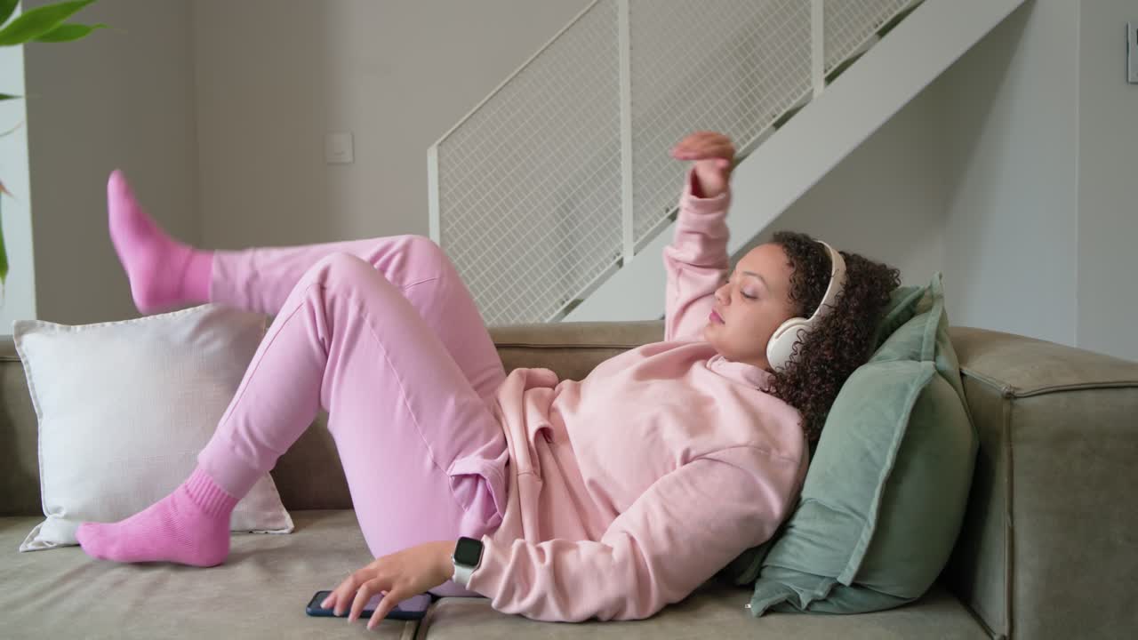 Donna afroamericana sdraiata sul divano con felpa rosa e cuffie, che scorre sul telefono e poi si rilassa