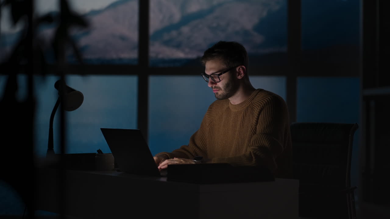 Young Guy Talking On Mobile Phone And Taking Notes Using Voice Assistant Writing In Notebook. Smiling Man Sitting At Desk With Laptop At Home Office Wearing Earbuds