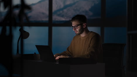 young guy talking on mobile phone and taking notes using voice assistant writing in notebook. smiling man sitting at desk with laptop at home office wearing earbuds