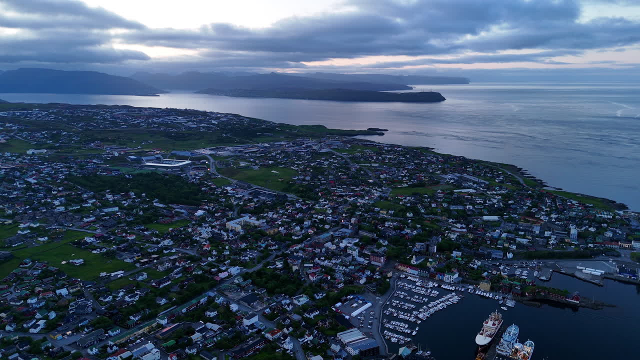 Cinematic aerial view of colorful village houses along a fjord in the Faroe Islands, surrounded by dramatic green mountains, black sand beach, and misty coastal scenery