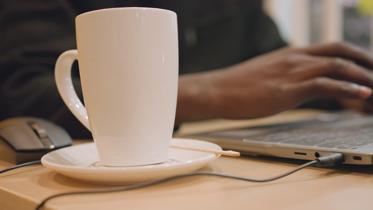 Tazza bianca in primo piano, mani nere che digitano vicino a laptop e mouse su una scrivania di legno, luce solare soffusa dalla finestra, scrittore che prende appunti con taccuino e penna, piattino e cucchiaino in ceramica visibili, atmosfera tranquilla.