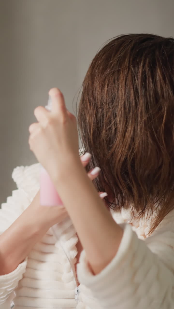 짧은 머리카락을 가진 여성은 화장실에서 화장품을 스프레이합니다. 부드러운 테리 로브를 입은 여자는 가운 욕실에서 머리카락에 유기농 제품을 뿌립니다.
