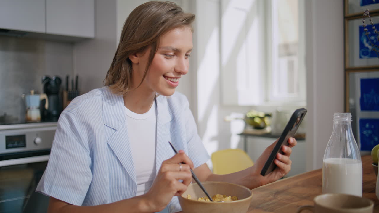 Relaxed girl eating cereal in sunny kitchen. Closeup home female looking mobile